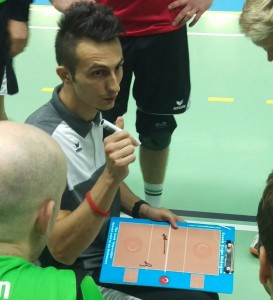 Das Foto zeigt einen Trainer mit einem Taktikboard während eines Volleyballspiels, auf dem er seinen Spielern letzte taktische Anweisungen gibt. 