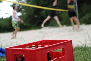 Das Bild zeigt die rote Sporttasche (eine Kiste Kölsch) vor dem Beachvolleyballfeld. Dort spielen gerade Volleyballer 2 gegen 2.