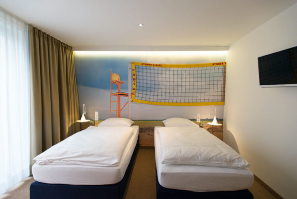 Das Foto zeigt ein Hotelzimmer im Volleyball Stil. An der Wand hängt ein Volleyballnetz und Schiedsrichterstuhl ist auf die Wand gemalt.