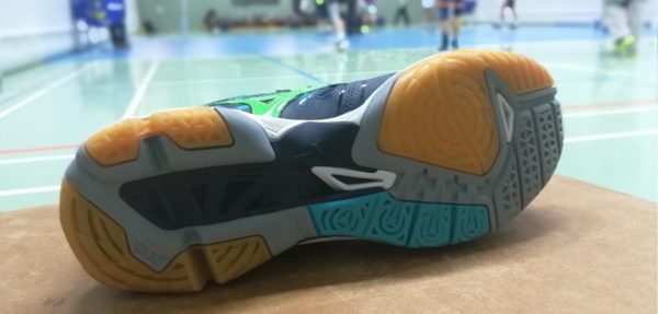 Das Foto zeigt die Sohle vom Volleyballschuh Sohle vom Mizuno Wave Tornado X2. Im Hintergrund sind unscharf Volleyballspieler beim Training zu sehen.