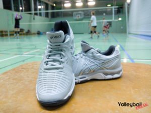 Die grauen Asics Netburner Ballistic Volleyballschuhpaar in der Frontansicht. Im Hintergrund trainieren Volleyballspielerinnen.