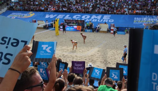 Auf dem Foto sieht man die Zuschauer die Klatschpappen ausgebreitet nach oben halten, während auf dem Center Court das Spiel weiterläuft.