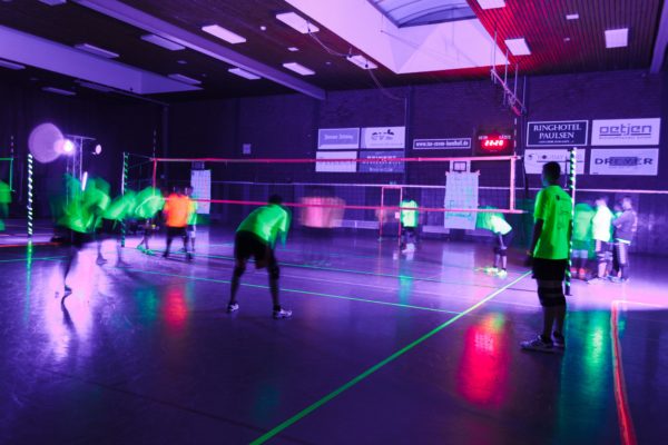 Auf dem Foto sieht man Spieler, Volleyballfeld und das Volleyballnetz beleuchtet von Schwarzlicht.